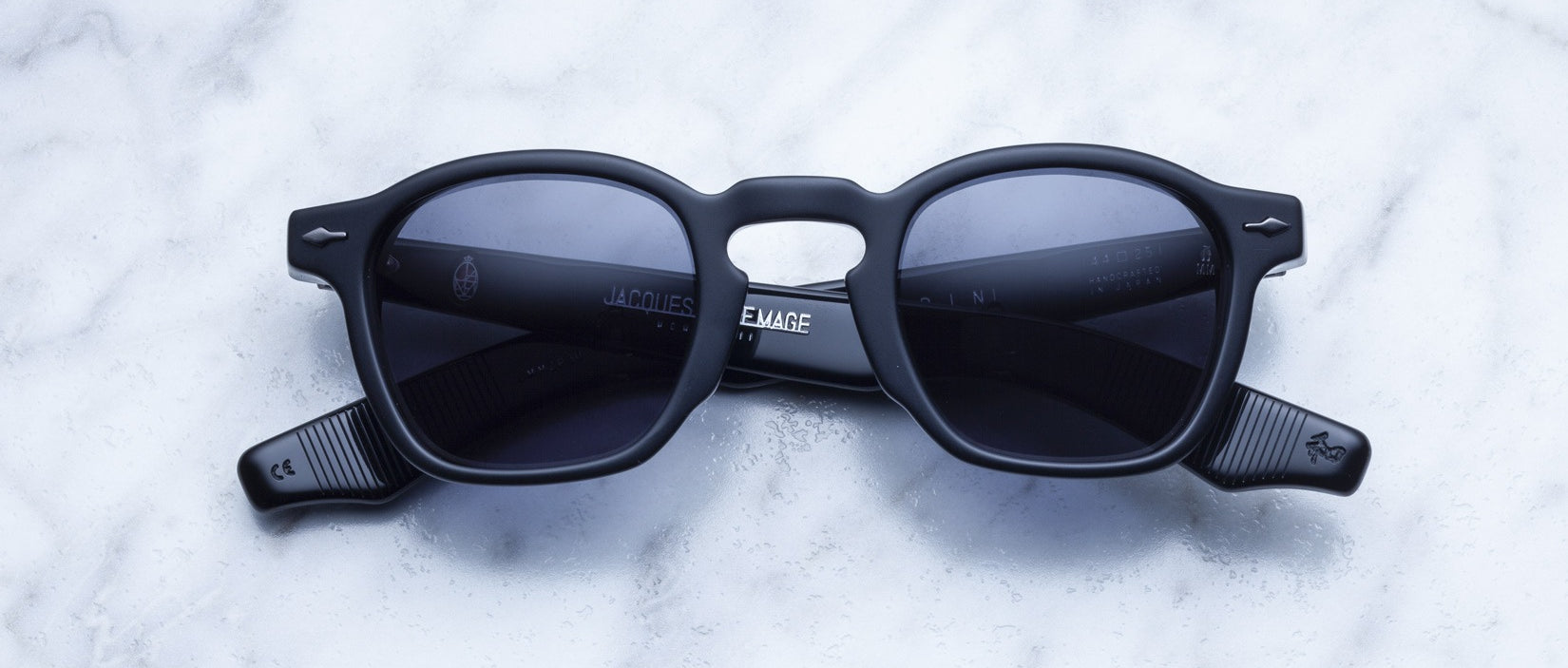 The Jacques Marie Mage Zephirin sunglasses by Jacques Marie Mage feature black handcrafted acetate frames and dark lenses, showcased on white marble. This limited edition eyewear epitomizes unique sophistication.