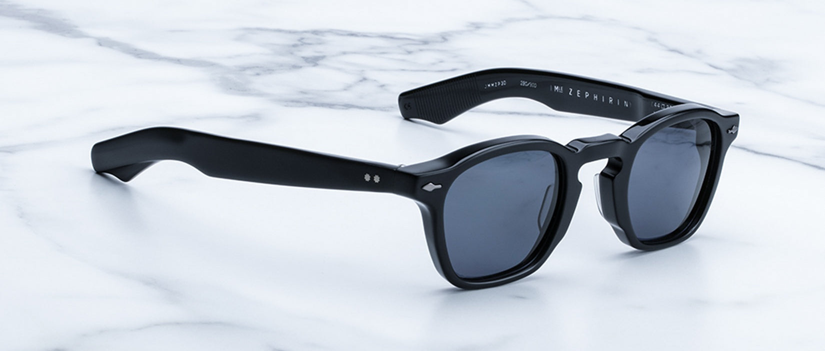 A pair of Jacques Marie Mage Zephirin black sunglasses with dark lenses and handcrafted acetate frames sits on a white marble surface.
