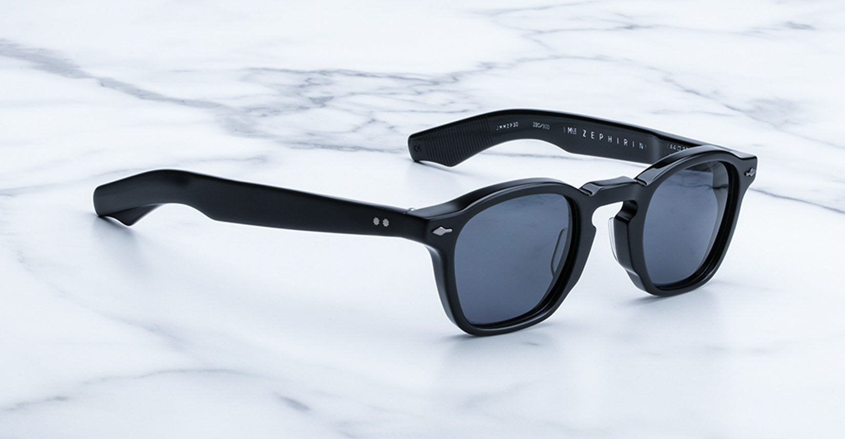 A pair of Jacques Marie Mage Zephirin black sunglasses with dark lenses and handcrafted acetate frames sits on a white marble surface.