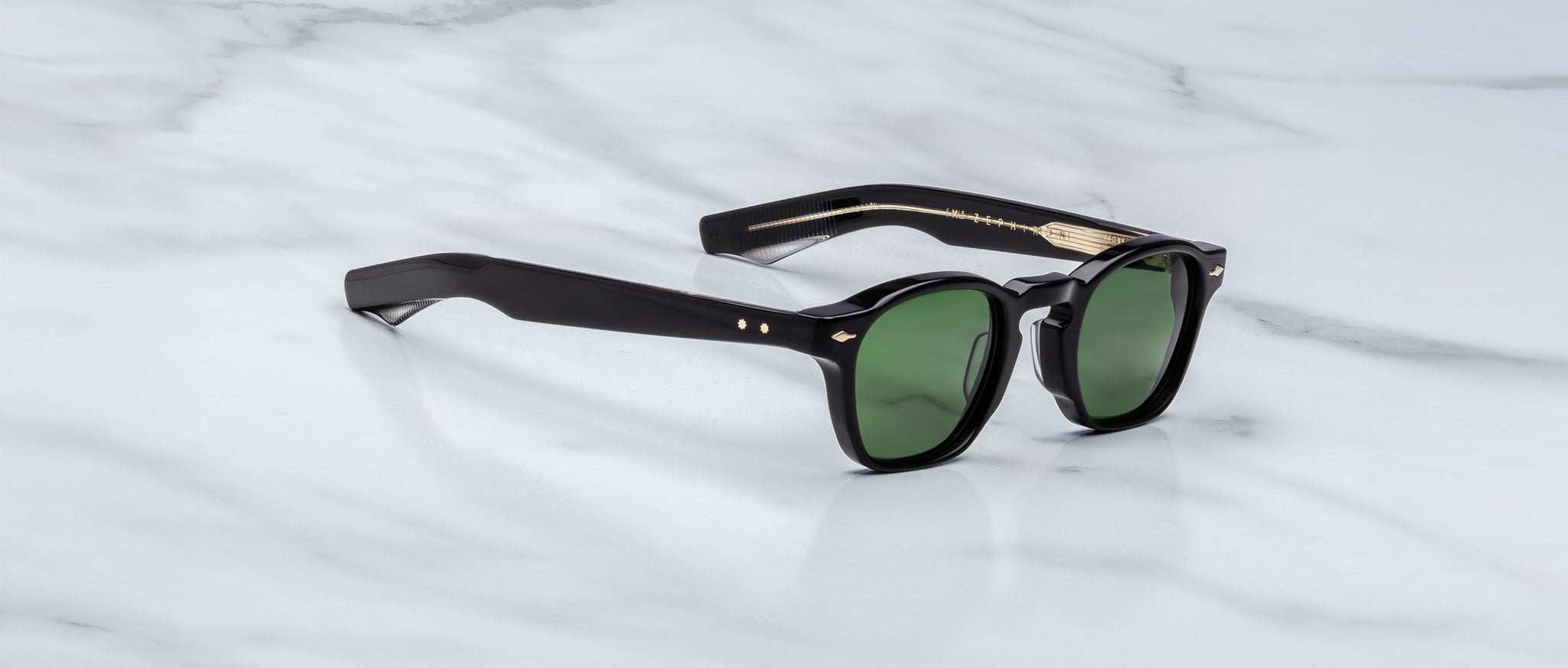 The Jacques Marie Mage Zephirin sunglasses feature green lenses and gold-detailed arms, showcased on a white marble surface, reflecting the elegance of limited edition Jacques Marie Mage handcrafted acetate eyewear.