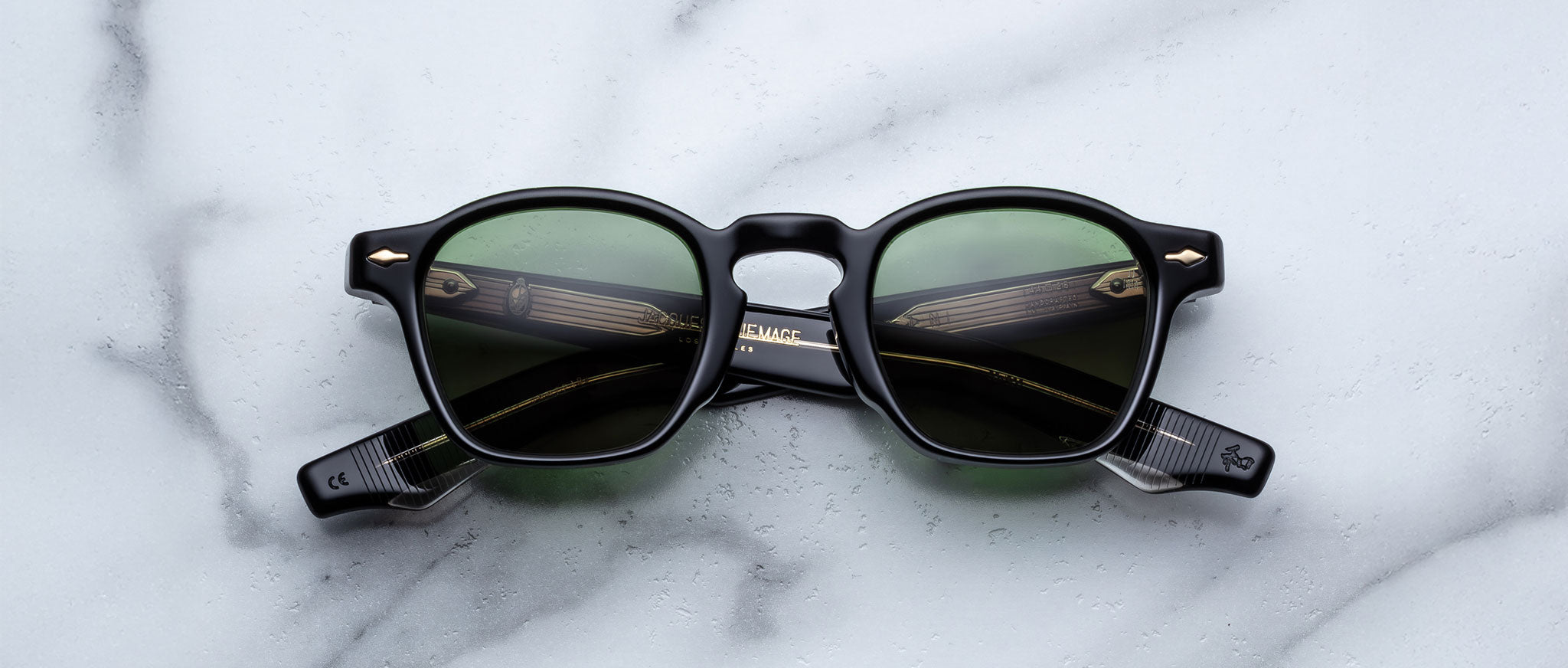 The Jacques Marie Mage Zephirin sunglasses by Jacques Marie Mage, featuring black round frames and green lenses reminiscent of handcrafted acetate, are displayed on a white marble surface.