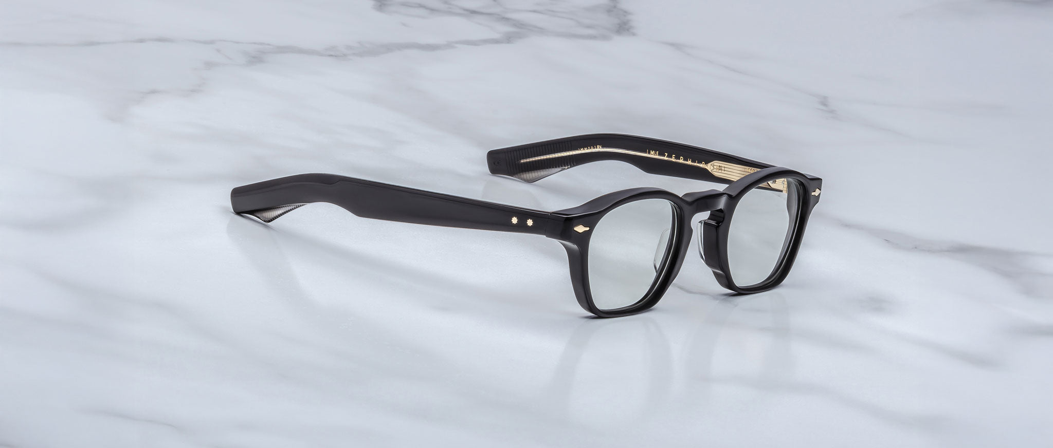Jacques Marie Mage Zephirin black-framed handcrafted acetate spectacles with clear lenses and gold accents by Jacques Marie Mage, displayed on a white marble surface.