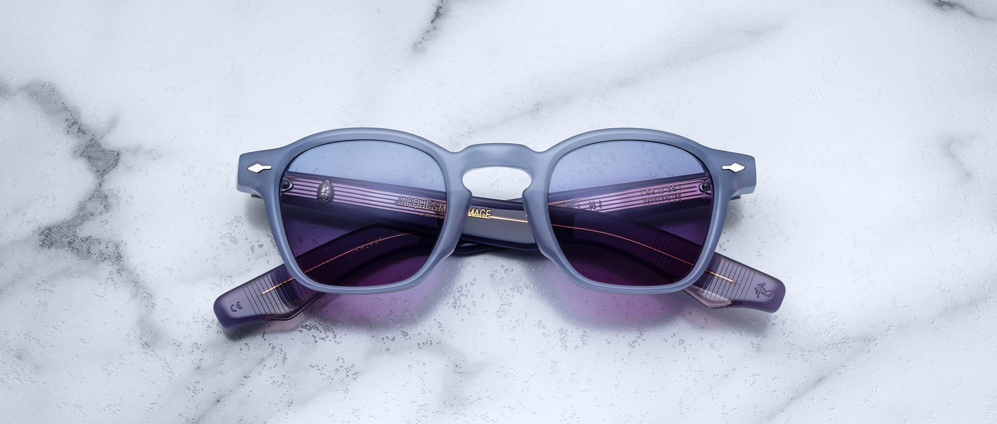 A pair of Jacques Marie Mage Zephirin sunglasses featuring square blue frames and purple-tinted lenses rests on a white marble surface, highlighting the brand’s exquisite handcrafted acetate eyewear.