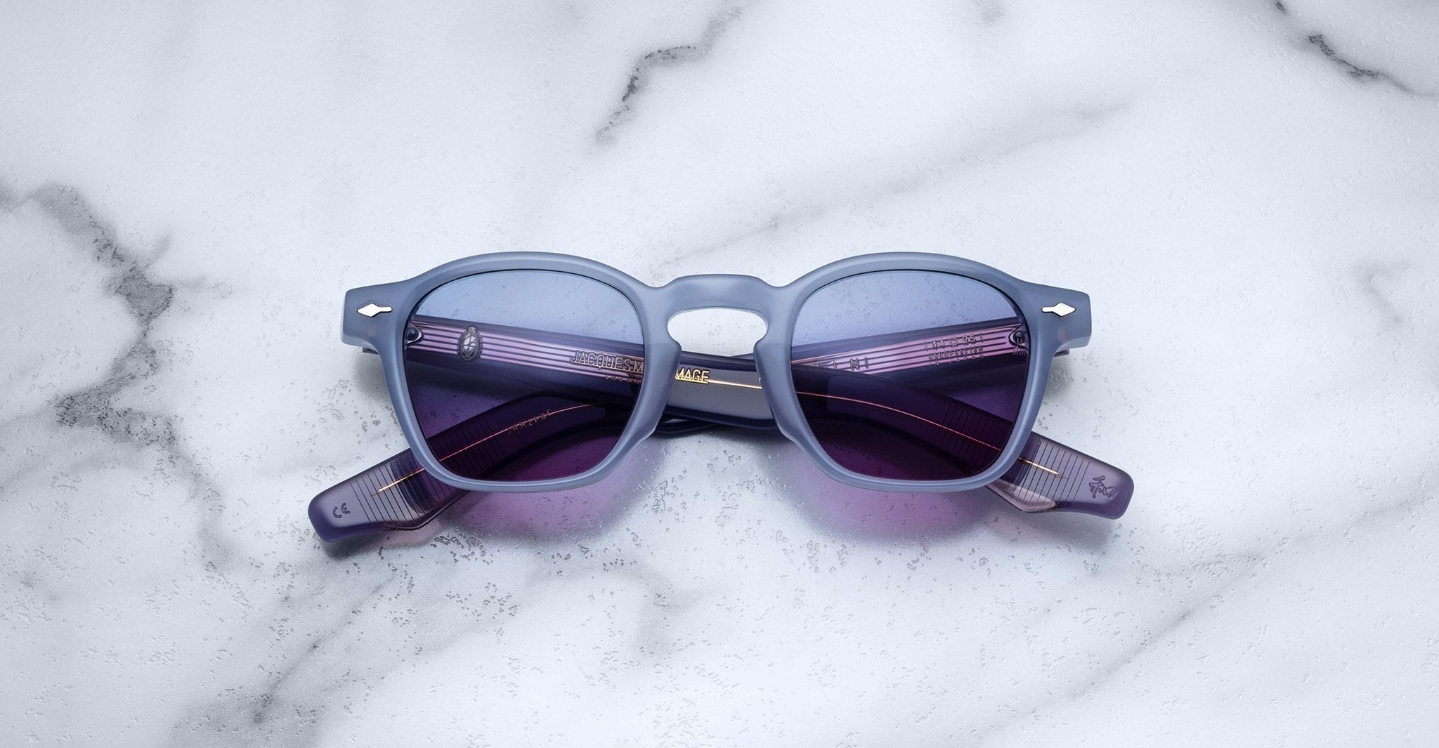 A pair of Jacques Marie Mage Zephirin sunglasses featuring square blue frames and purple-tinted lenses rests on a white marble surface, highlighting the brand’s exquisite handcrafted acetate eyewear.