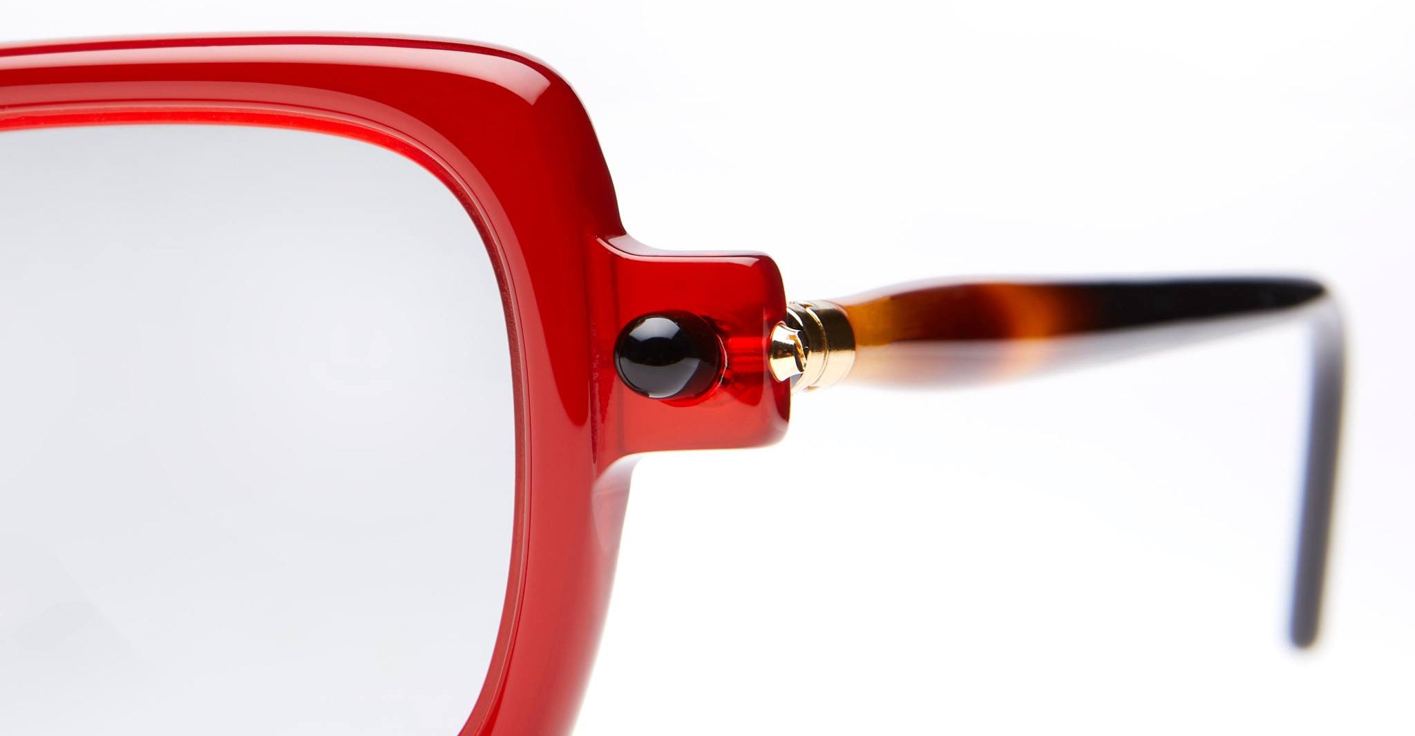 Close-up of the left side of a pair of eyeglasses with a thick, glossy red frame, clear lens, black hinge detail, and tortoiseshell-patterned temple against a white background.