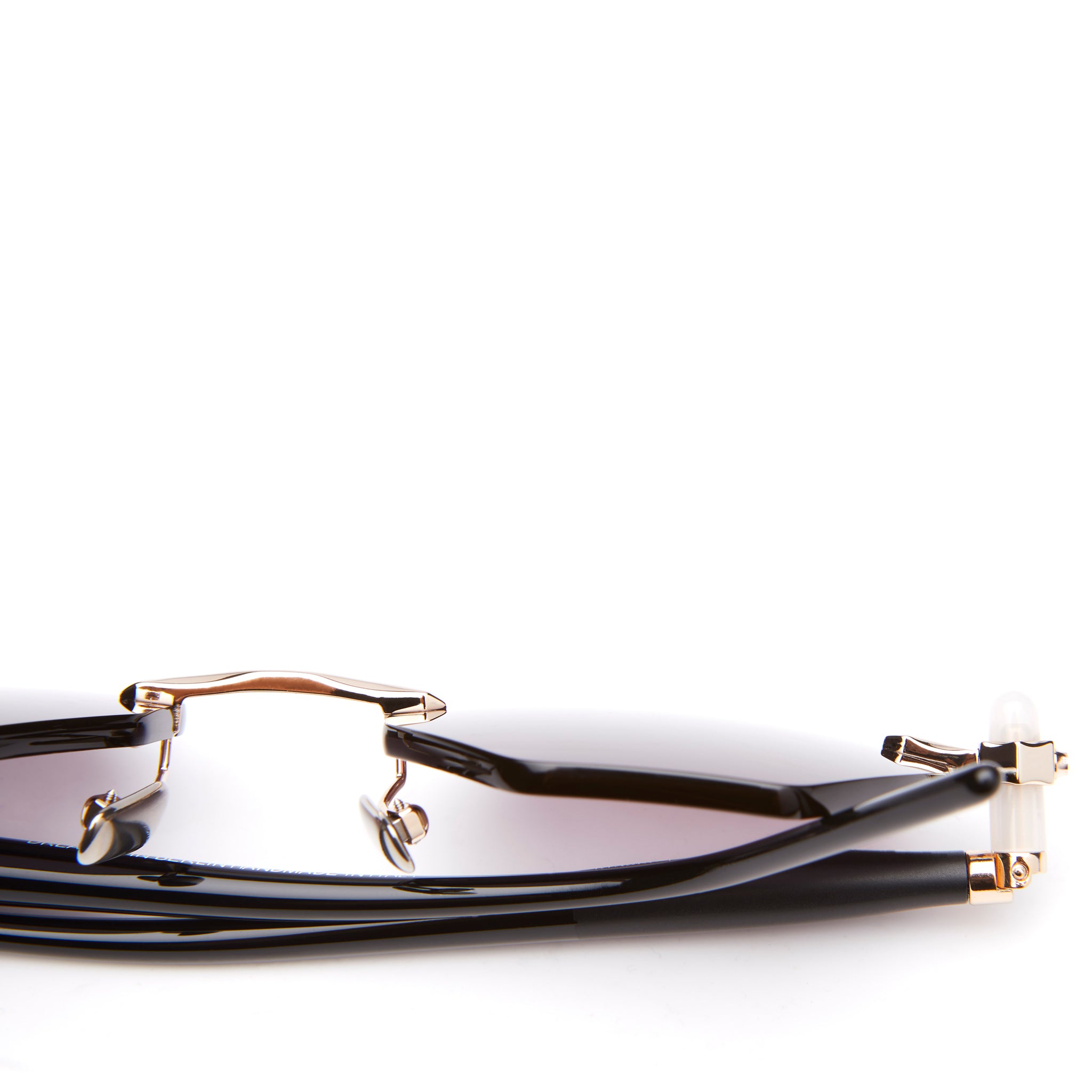 A close-up of black sunglasses with gold metal bridge and accents, lying folded against a white background. The gradient lenses transition from dark to light.