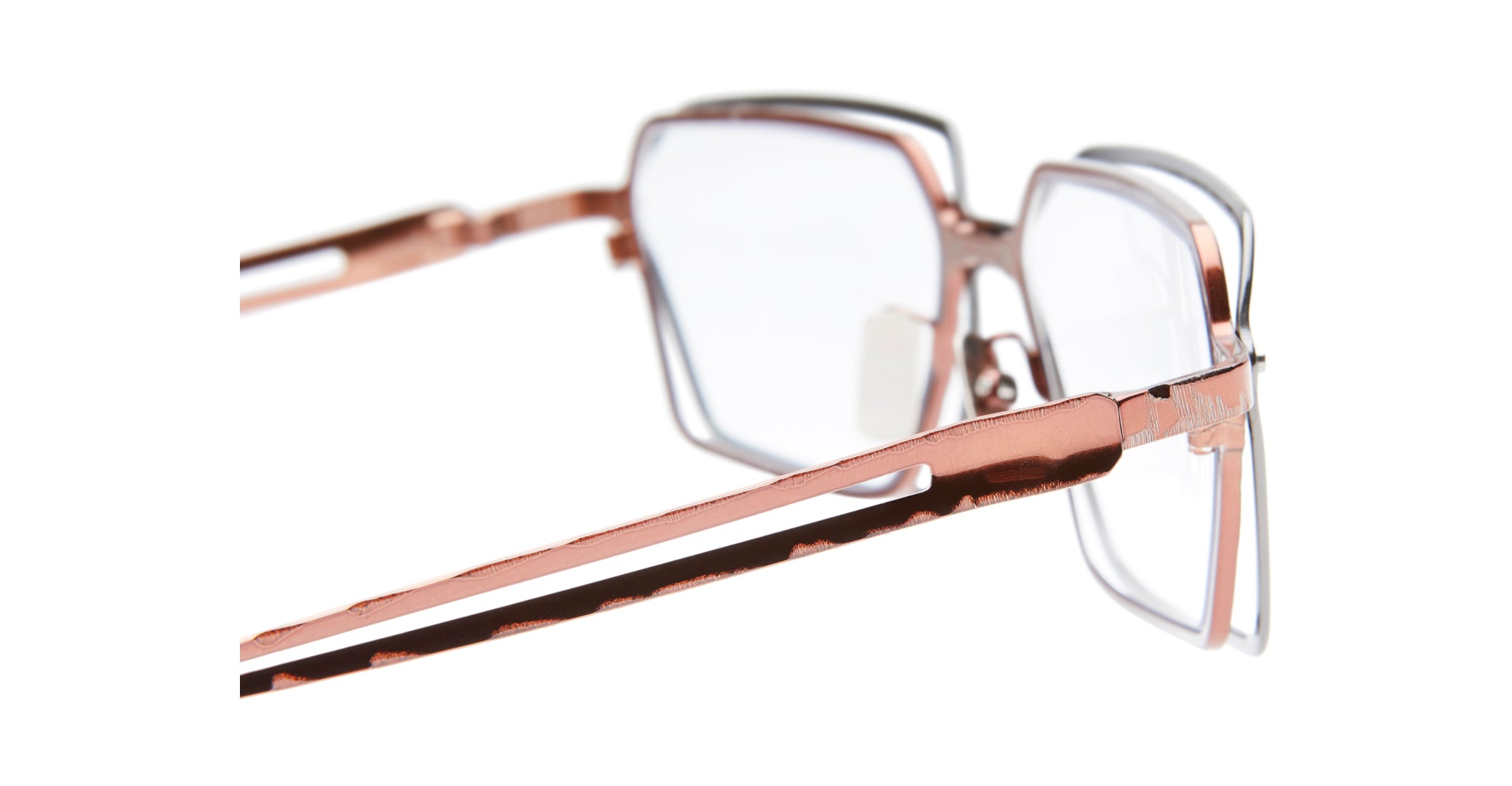 Close-up of Kuboraum H15 eyeglasses with rose gold metallic frames and clear lenses, shown from the side. The white background highlights their geometric design and shiny finish.