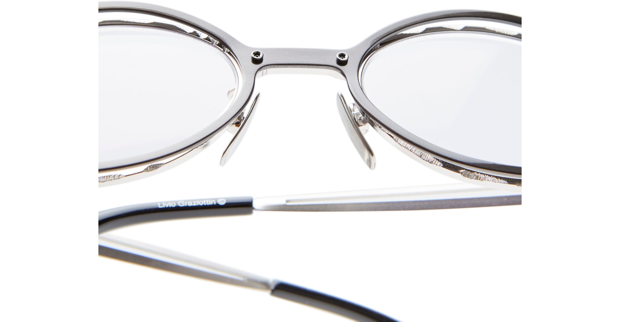 Close-up of Kuboraum H18 silver metal eyeglasses with clear lenses, highlighting the nose pads and bridge. The black temple arm appears slightly out of focus in the foreground.
