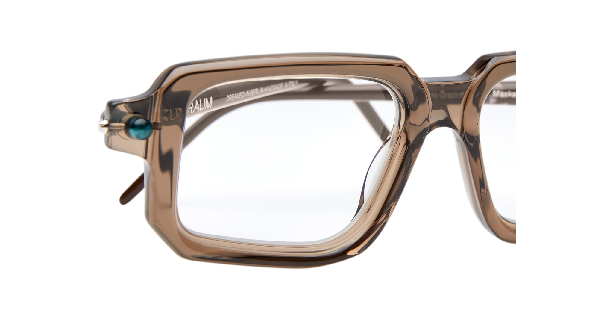 Close-up of the Kuboraum P23 eyeglass frames, featuring a rectangular shape, transparent brown color, clear lenses, and a small blue accent near the left hinge.