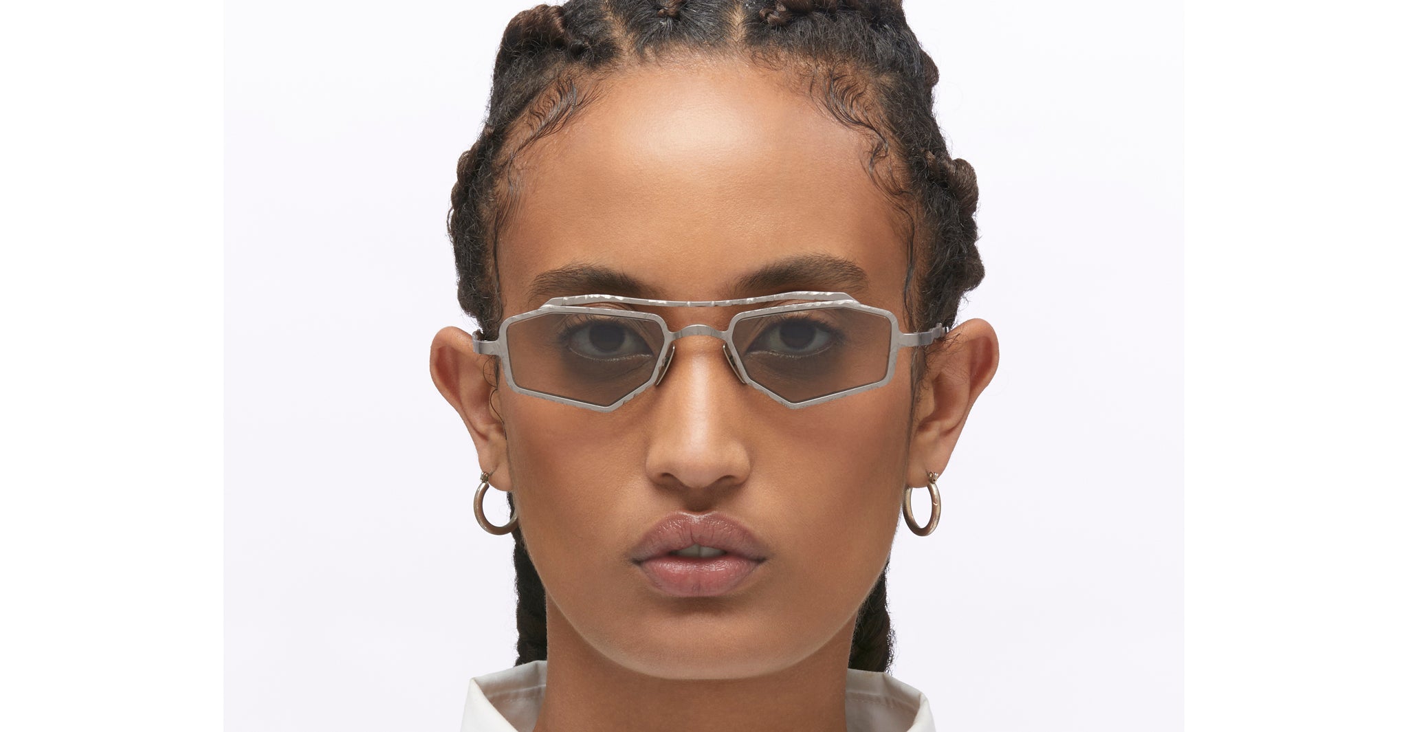 A person with braided hair wears Kuboraum Z23 geometric, silver-rimmed sunglasses and hoop earrings, gazing neutrally at the camera against a plain white background.