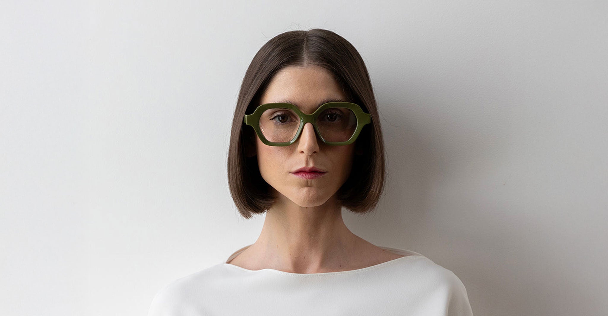 A person with straight, chin-length brown hair wears Lapima's Alice large green geometric glasses and a white top, standing against a plain white background.