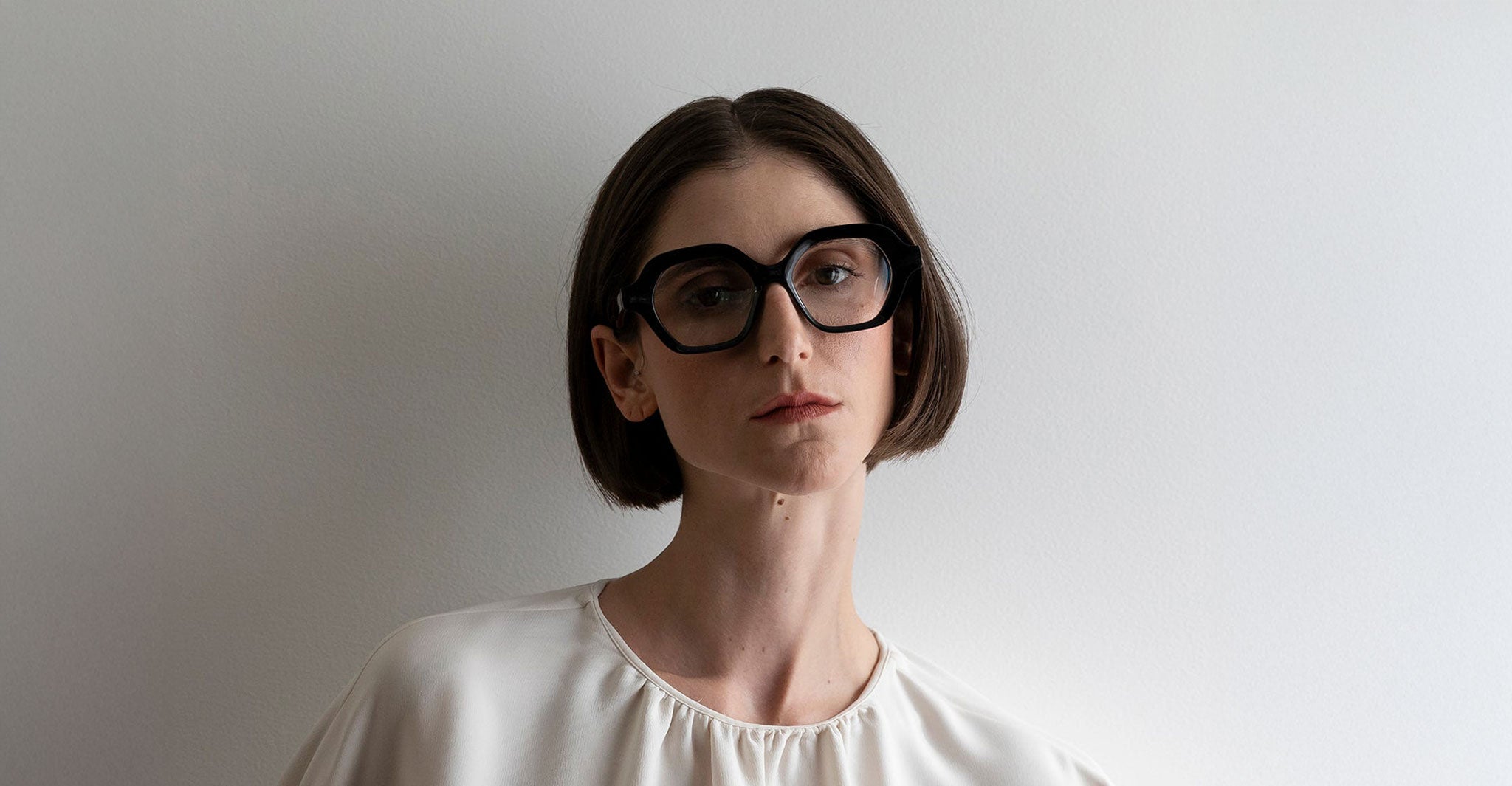 A person with short brown hair, wearing Lapima Alice eyeglasses and a white blouse, stands against a plain white background, facing the camera with a neutral expression.