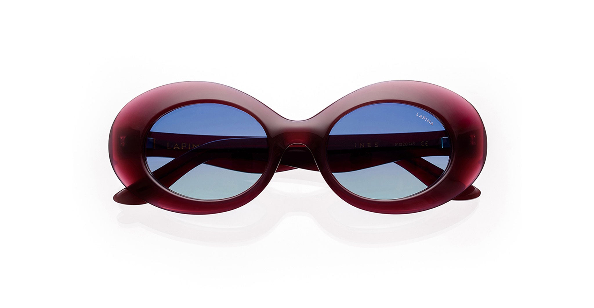 The Lapima Ines sunglasses feature thick, oval-shaped maroon frames and blue-tinted lenses, displayed against a white background.