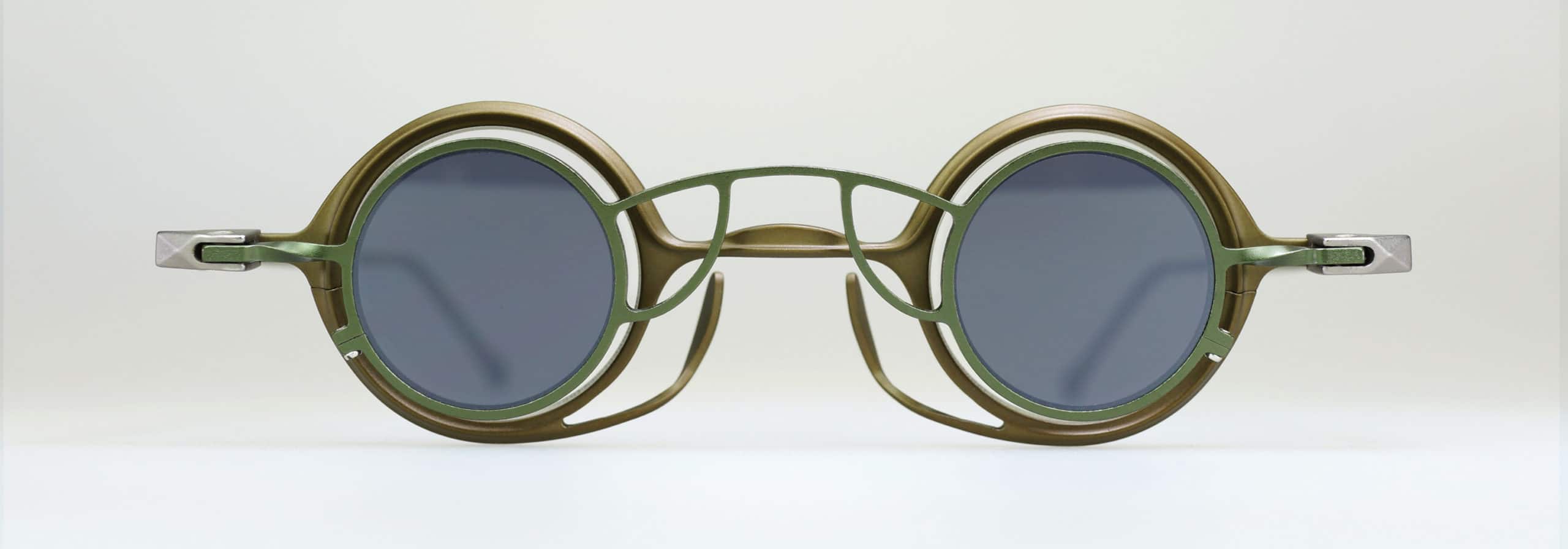 Round sunglasses with olive green metal frames, dark lenses, and a unique double bridge design with angular geometric details, displayed against a plain light background.