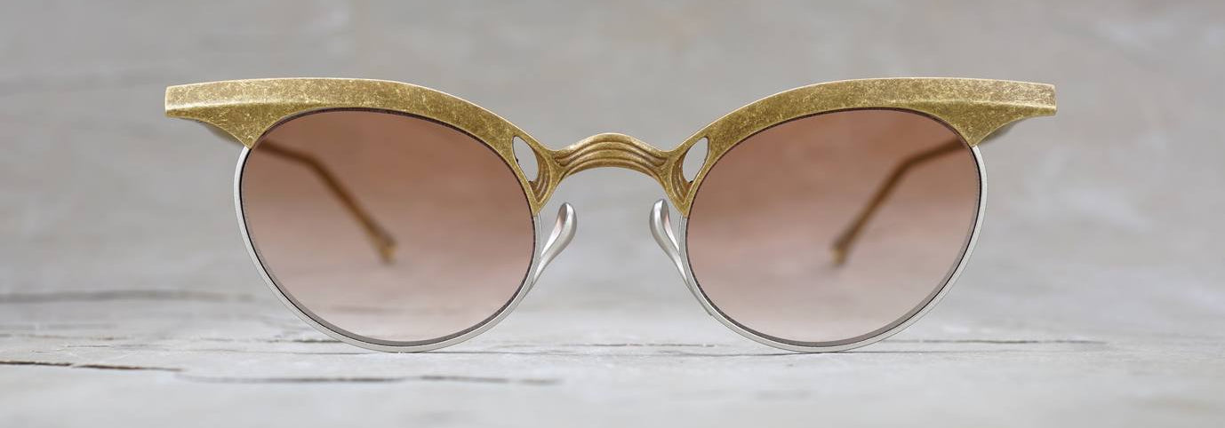 Round sunglasses with gold textured frames, silver rims, and brown gradient lenses placed on a light gray surface, photographed straight on with a blurred background.