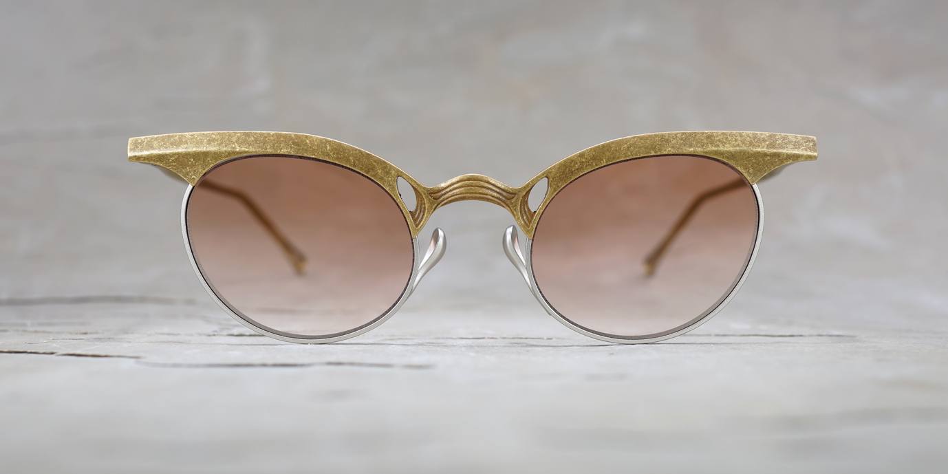 Round sunglasses with gold textured frames, silver rims, and brown gradient lenses placed on a light gray surface, photographed straight on with a blurred background.