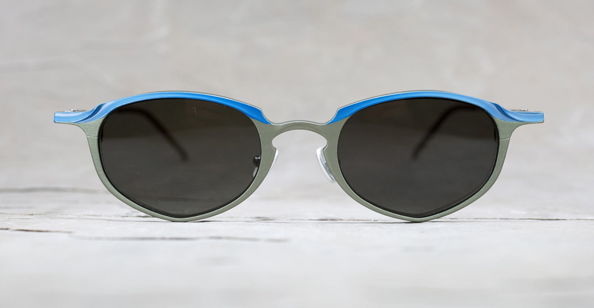 A pair of round sunglasses with dark lenses, a metallic gray frame, and a bright blue accent along the top edge, displayed against a light, neutral background.