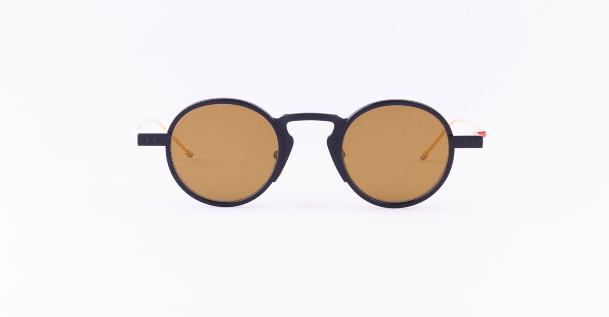 The Thom Browne 931 sunglasses feature round black frames, brown-tinted lenses, and thin arms with orange-tinted tips, displayed centered on a plain white background.