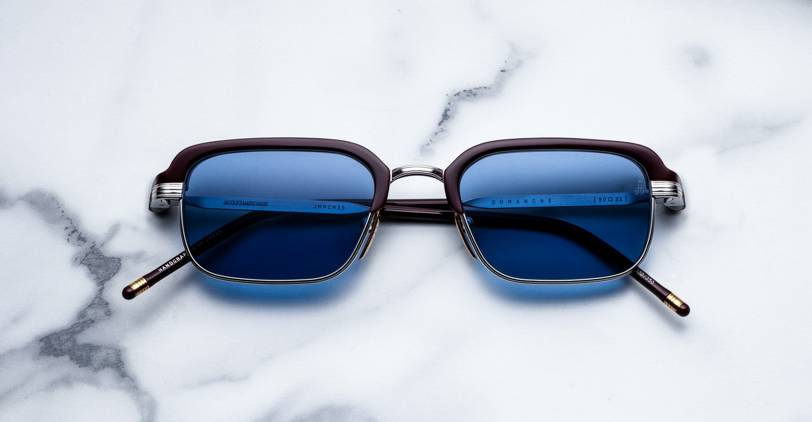 A pair of Jacques Marie Mage Comanche square eyeglasses with blue-tinted lenses, metal rims, and brown temples sits on a white marble surface.