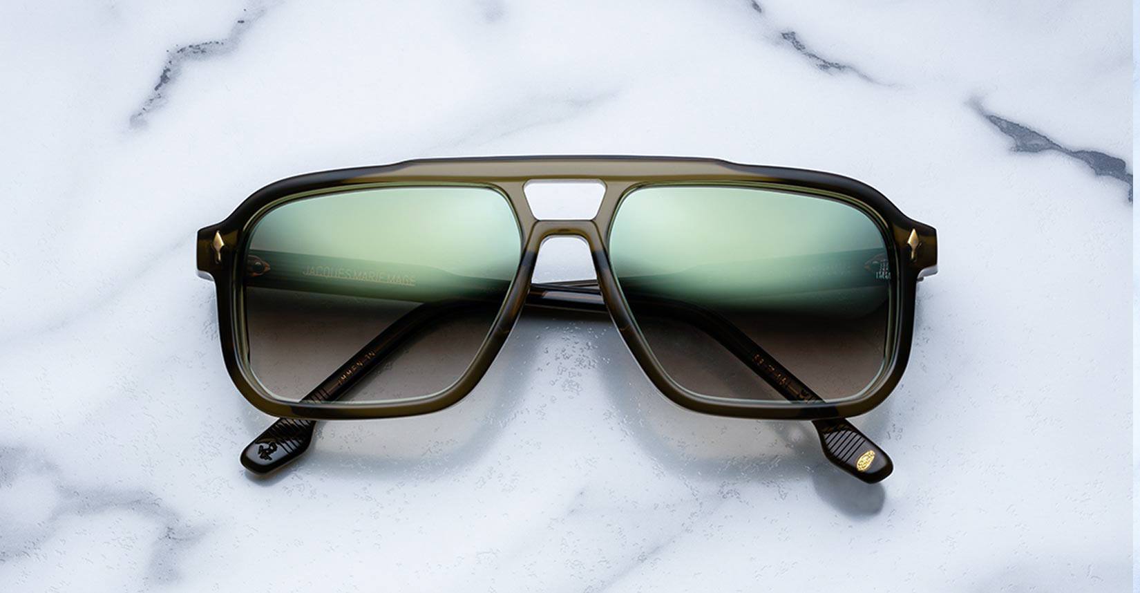 A pair of olive-green sunglasses with large, rectangular gradient lenses rests on a white marble surface with gray veining.