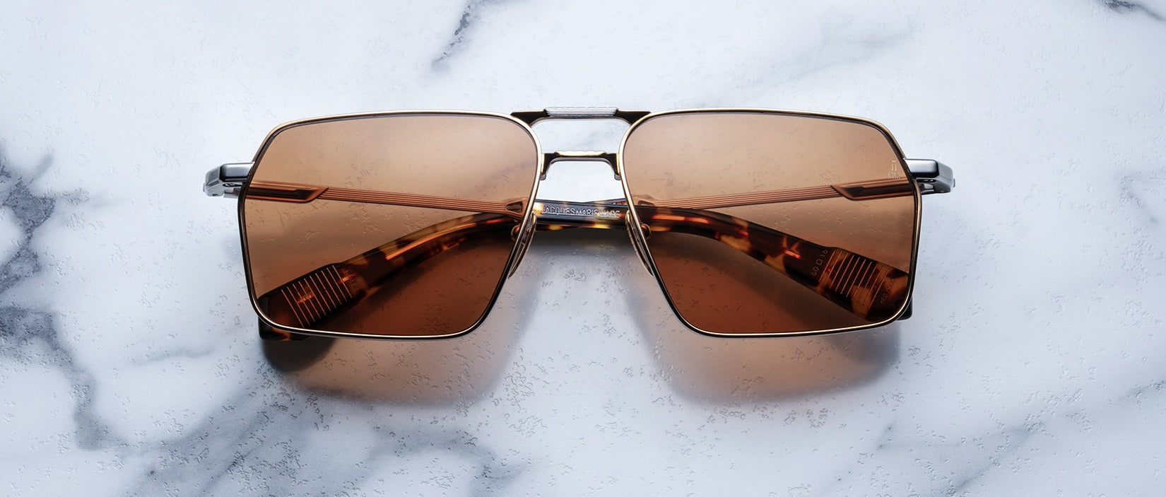 The Jacques Marie Mage GT features brown lenses and a square metallic frame, showcased on a white marble surface. These sunglasses offer a stylish, modern design.