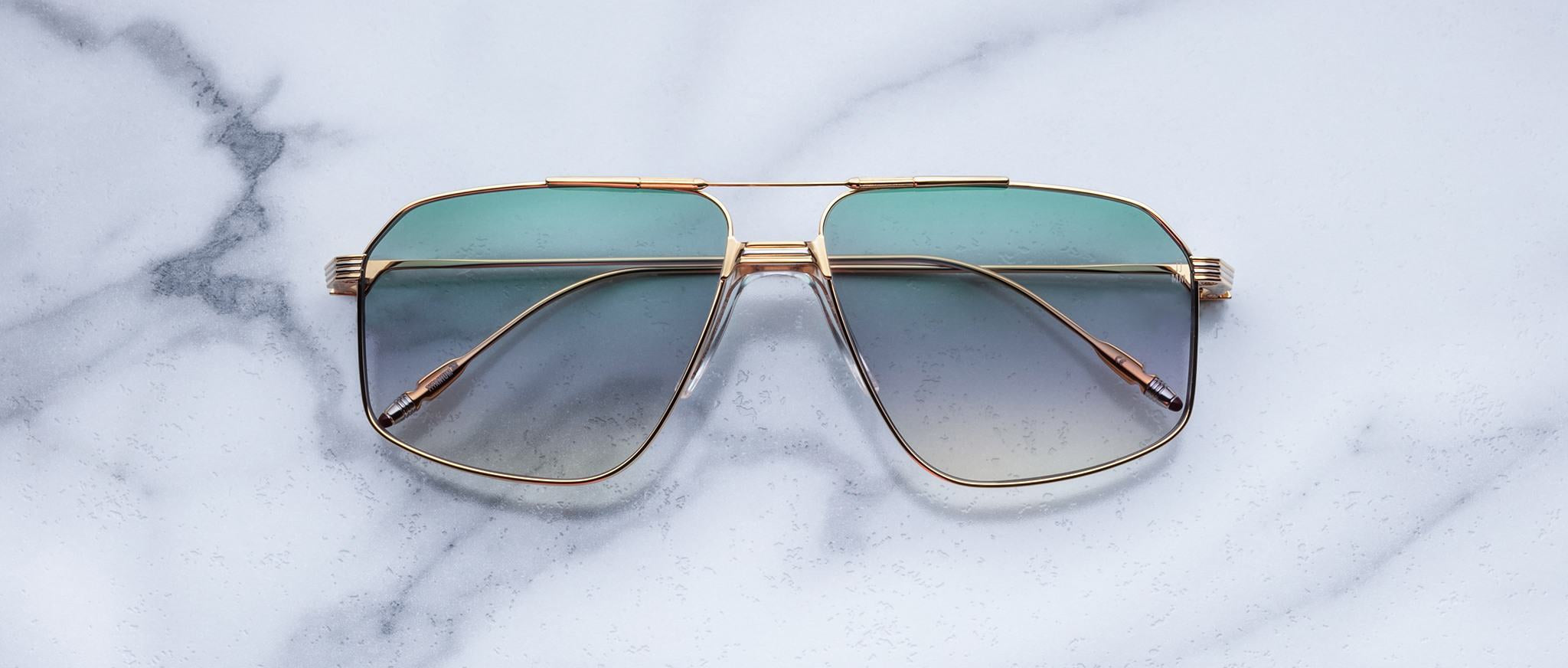 Jacques Marie Mage Jagger sunglasses by Jacques Marie Mage, featuring gold-tone oversized aviator frames with green gradient lenses, displayed elegantly on a white marble surface.