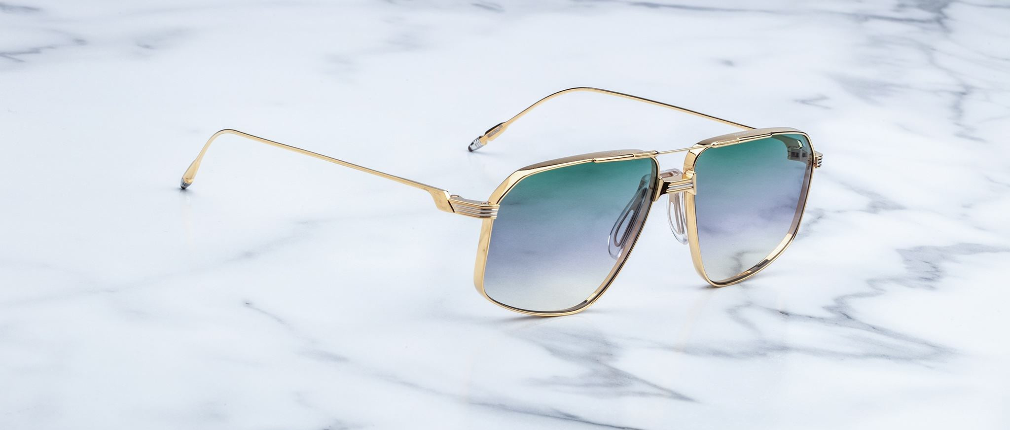 Jacques Marie Mage Jagger oversized aviator sunglasses, featuring gold frames and green gradient lenses, from Jacques Marie Mage, rest on a sleek white marble surface.
