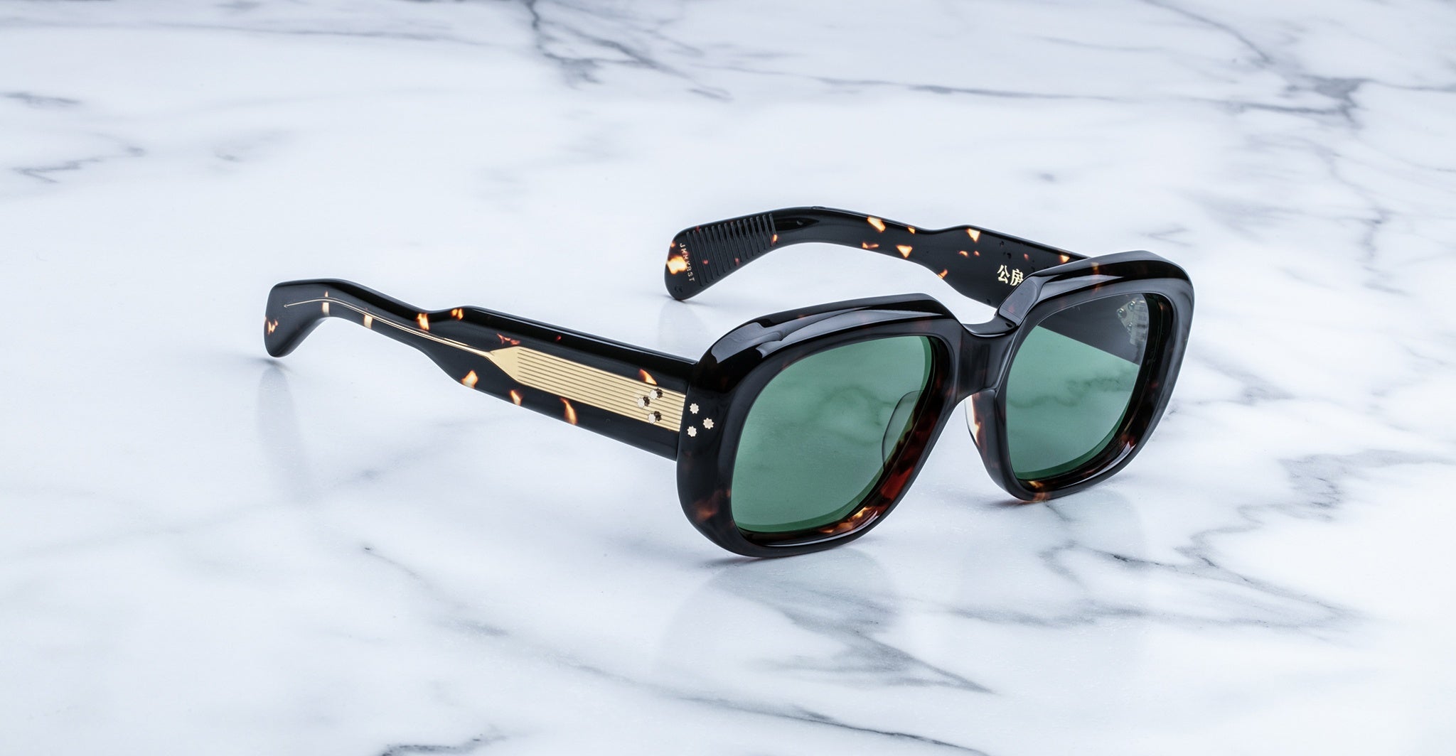 A pair of tortoiseshell sunglasses with thick frames and green lenses, featuring gold accents on the arms, displayed on a white marble surface.