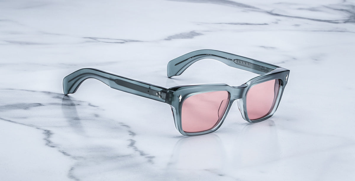 The Jacques Marie Mage Molino sunglasses in Hunter feature square, thick, translucent gray frames and pink-tinted lenses, displayed on a white marble surface with gray veining.
