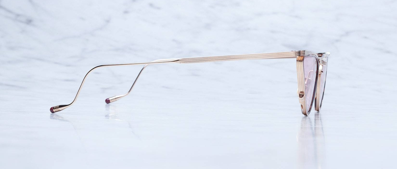 A pair of stylish gold-rimmed eyeglasses shown from the side, resting on a reflective marble surface with a light, cloudy background.