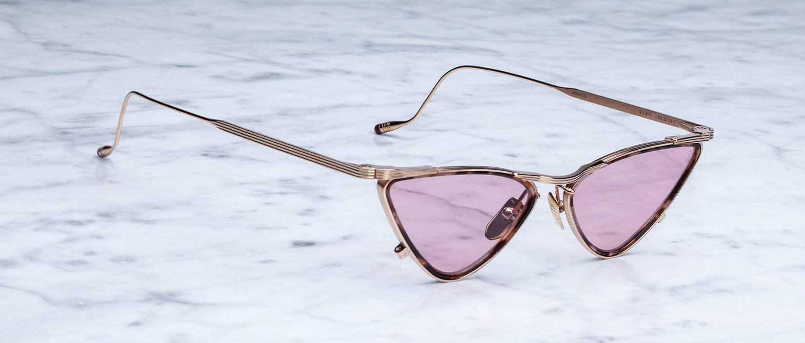 Rose gold cat-eye sunglasses with pink-tinted lenses are placed on a white marble surface. The frames have a modern, angular design and thin metal arms.
