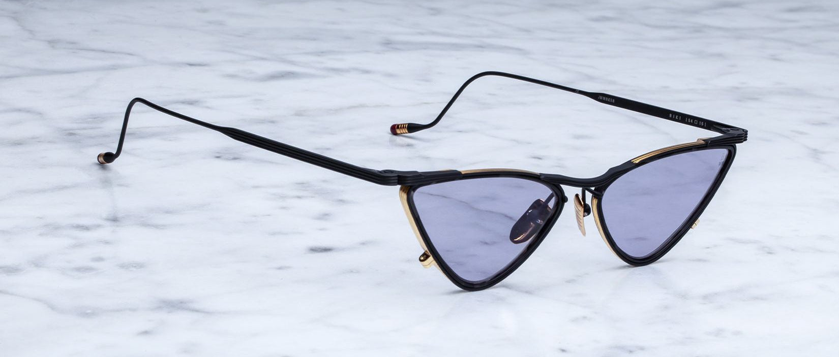 A pair of black and gold cat-eye sunglasses with purple-tinted lenses sits on a light gray marble surface.
