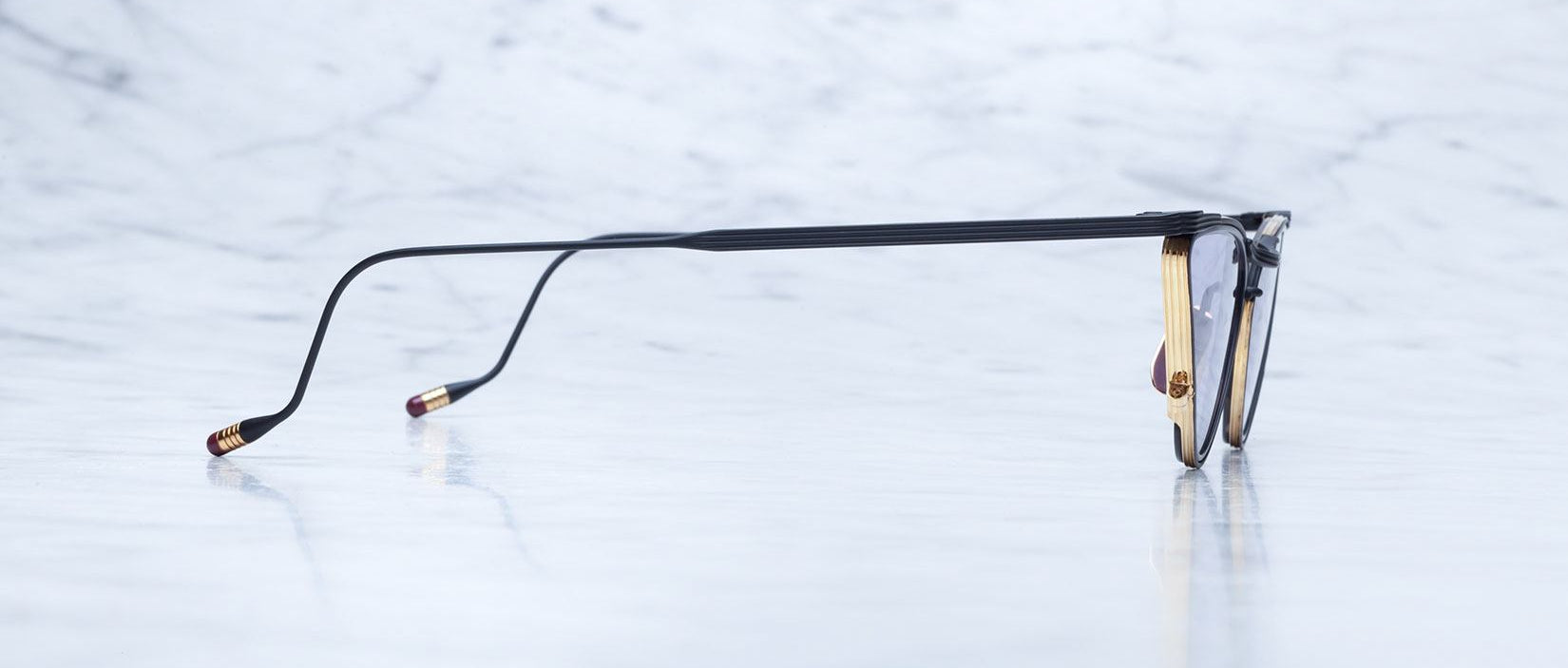 A pair of eyeglasses viewed from the side, showing thin black arms with gold accents on the temple tips, resting on a white marble surface with a light gray background.