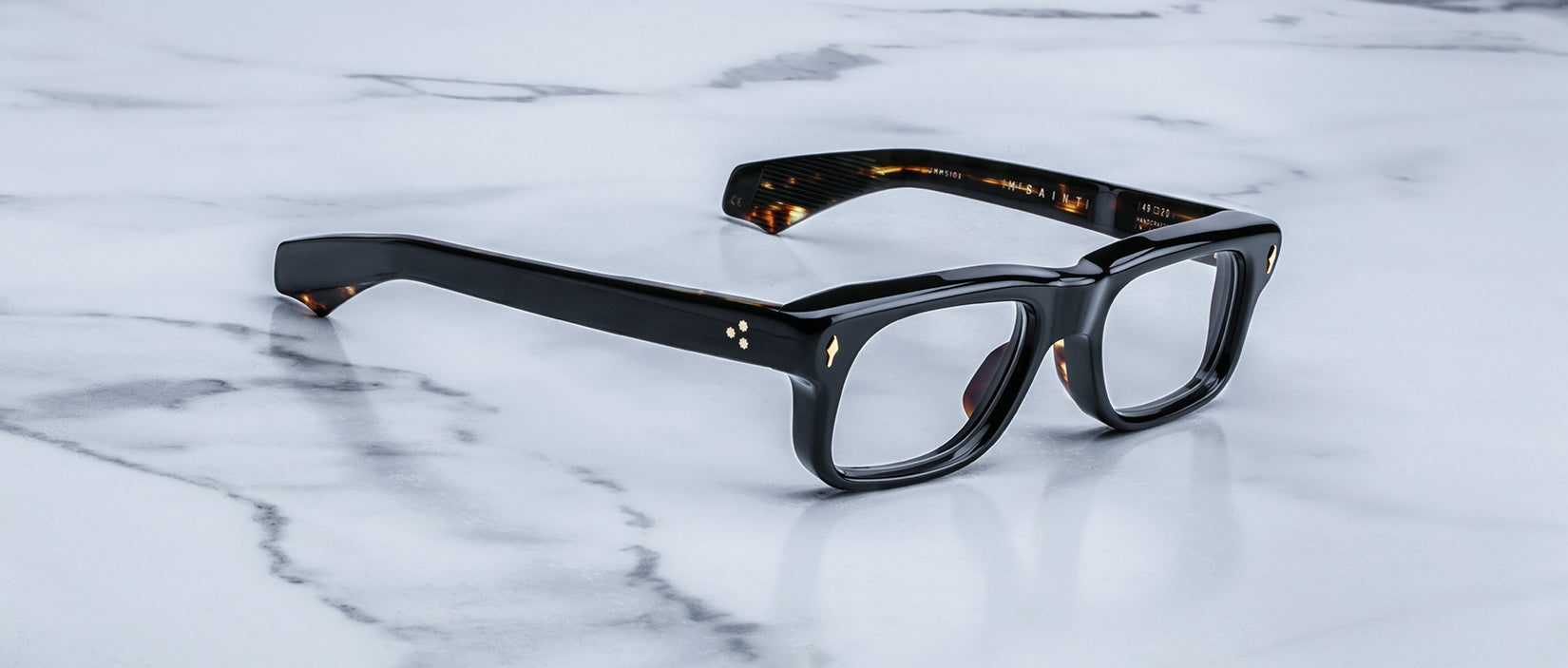 Jacques Marie Mage Saint eyeglasses with thick black rims and gold accents, front view, eyewear placed on a flat surface.