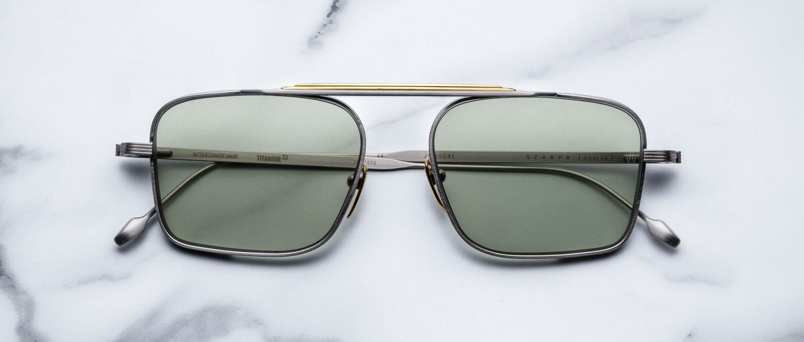 The Jacques Marie Mage Scarpa sunglasses feature green-tinted rectangular lenses, a slim metallic frame, and a gold bar across the top, shown on a white marble surface.
