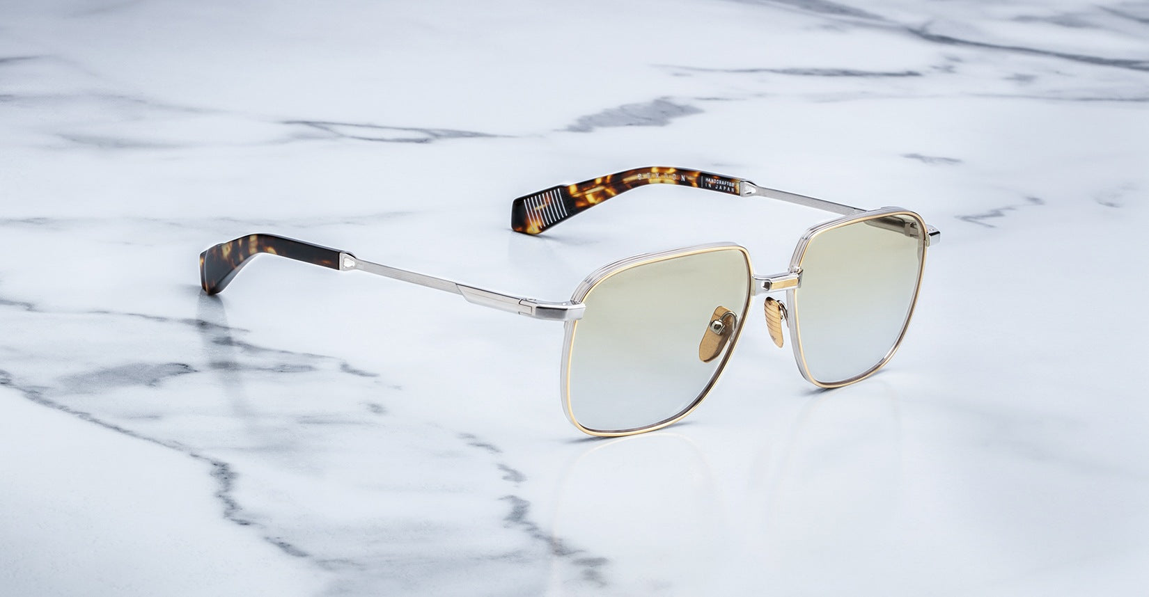 A pair of stylish sunglasses with gold metal frames and light tinted lenses rests on a smooth white marble surface. The temples have a distinctive brown tortoiseshell pattern at the ends.