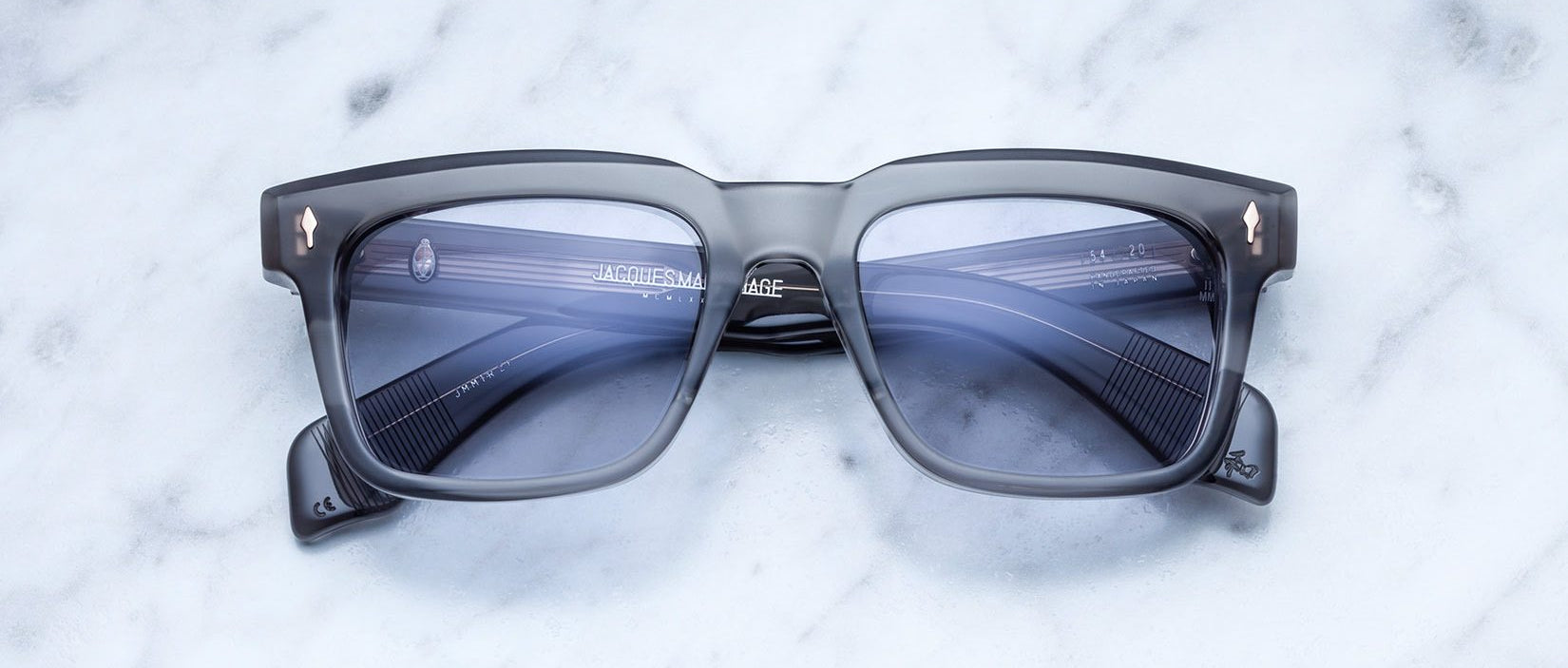 The Jacques Marie Mage Torino sunglasses by Jacques Marie Mage feature bold rectangular black frames with blue tinted lenses, displayed on a white marble surface.