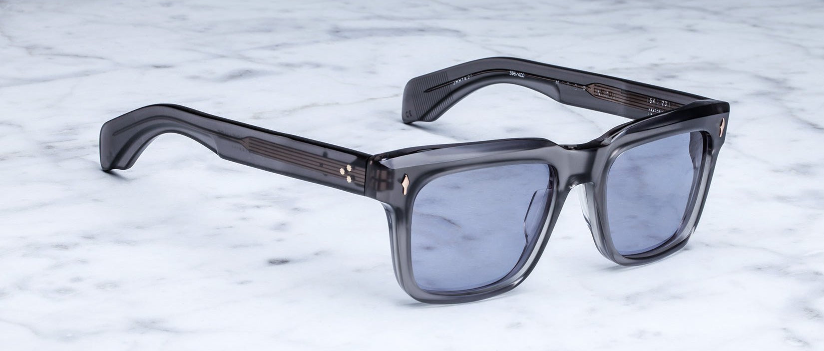 The Jacques Marie Mage Torino, bold rectangular frames in gray translucent acetate with blue-tinted lenses, rest elegantly on a white marble surface.