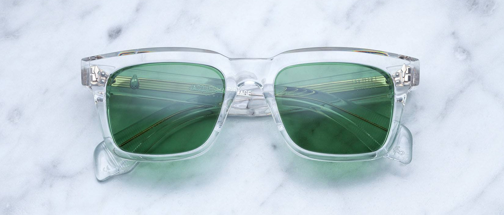 The Jacques Marie Mage Torino sunglasses by Jacques Marie Mage feature a clear frame and green tinted lenses, inspired by bold rectangular eyeglass designs, and rest elegantly on a white marble surface—a tribute to handcrafted acetate eyewear.