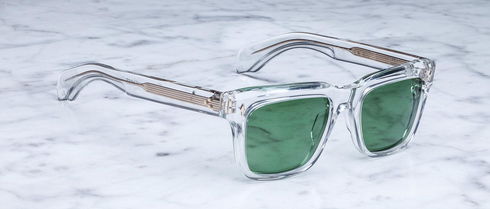 Jacques Marie Mage Torino sunglasses by Jacques Marie Mage feature clear frames and green tinted lenses, resting on white marble and highlighting their bold rectangular shape and exquisite handcrafted acetate craftsmanship.