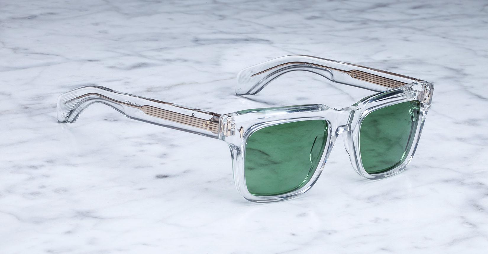 Jacques Marie Mage Torino sunglasses by Jacques Marie Mage feature clear frames and green tinted lenses, resting on white marble and highlighting their bold rectangular shape and exquisite handcrafted acetate craftsmanship.