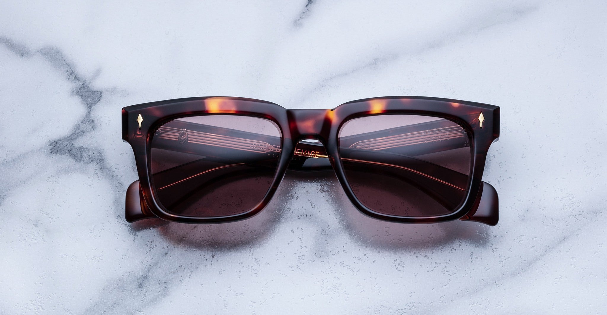 Jacques Marie Mage Torino sunglasses with bold square tortoiseshell frames and dark lenses rest on a white marble surface, showcasing the handcrafted acetate style of Jacques Marie Mage.