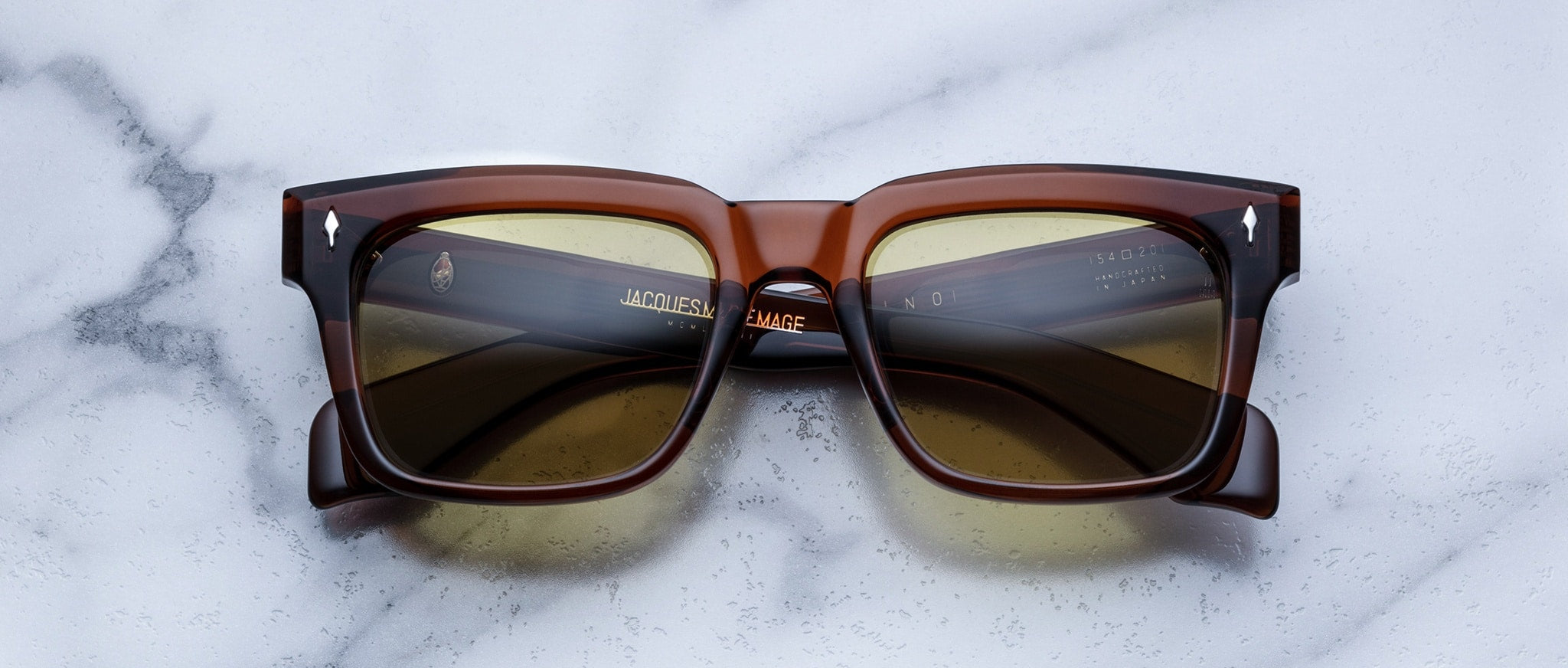 A pair of Jacques Marie Mage Torino rectangular brown sunglasses with dark lenses and bold handcrafted acetate frames rests on a white marble surface.