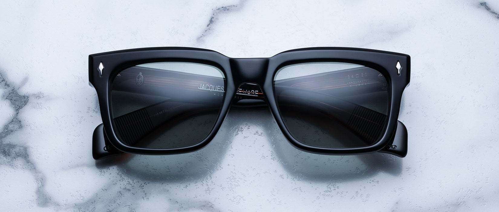The Jacques Marie Mage Torino, bold black rectangular eyeglass frames with dark lenses, rest on a white marble surface, highlighting the brand's handcrafted acetate artistry.