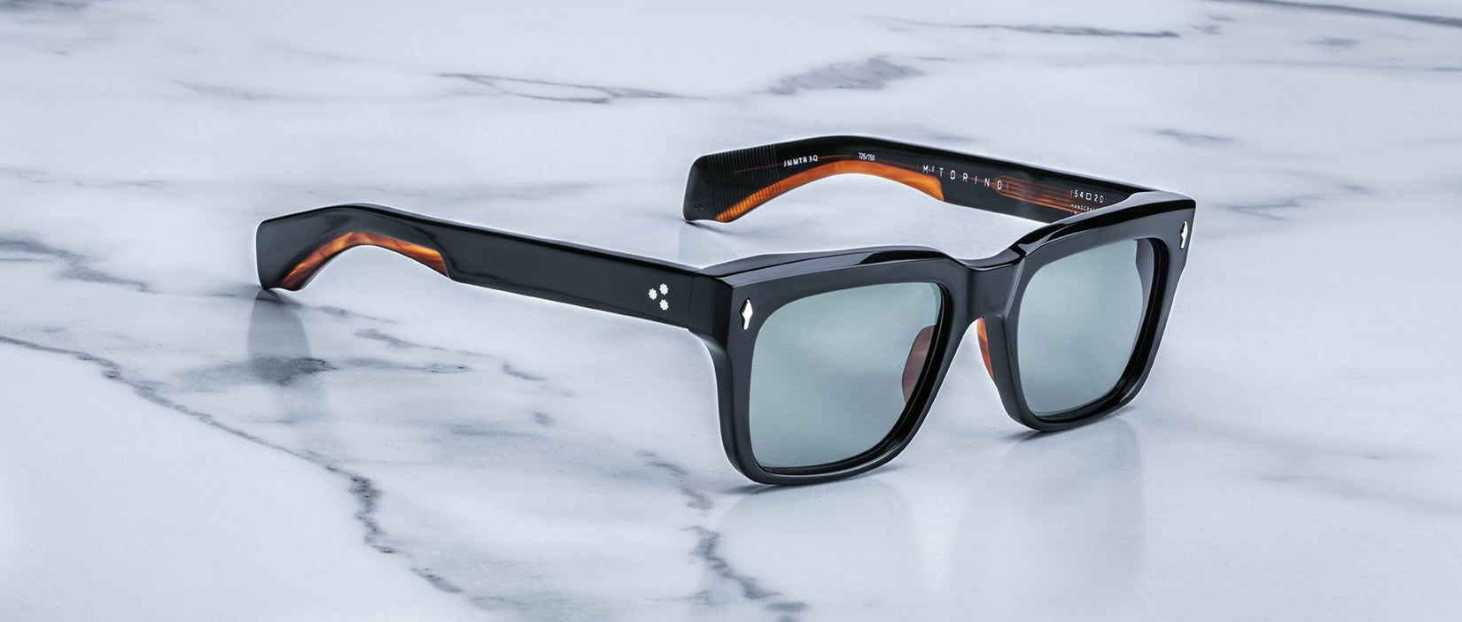 The Jacques Marie Mage Torino sunglasses—black rectangular frames with dark lenses and brown inner arms—crafted from premium acetate, rest stylishly on a white marble surface.