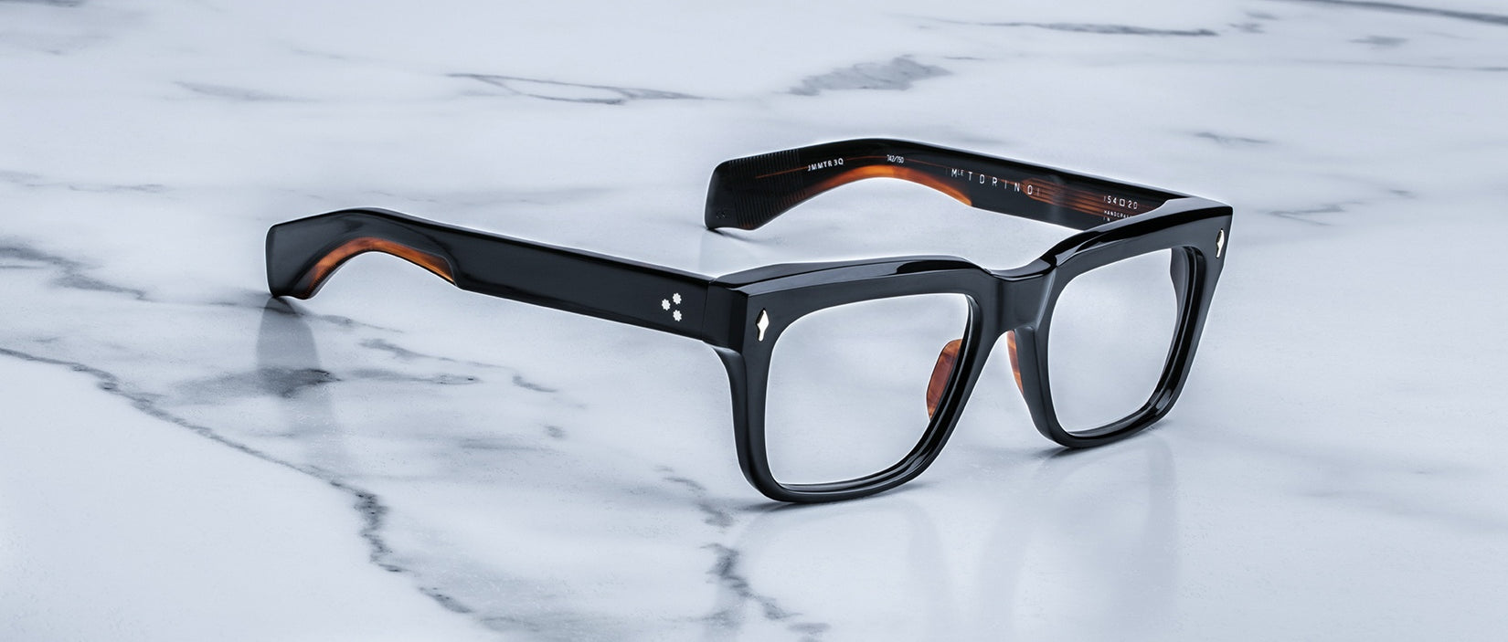 A pair of Jacques Marie Mage Torino rectangular eyeglass frames in black handcrafted acetate by Jacques Marie Mage rests on a white marble surface.
