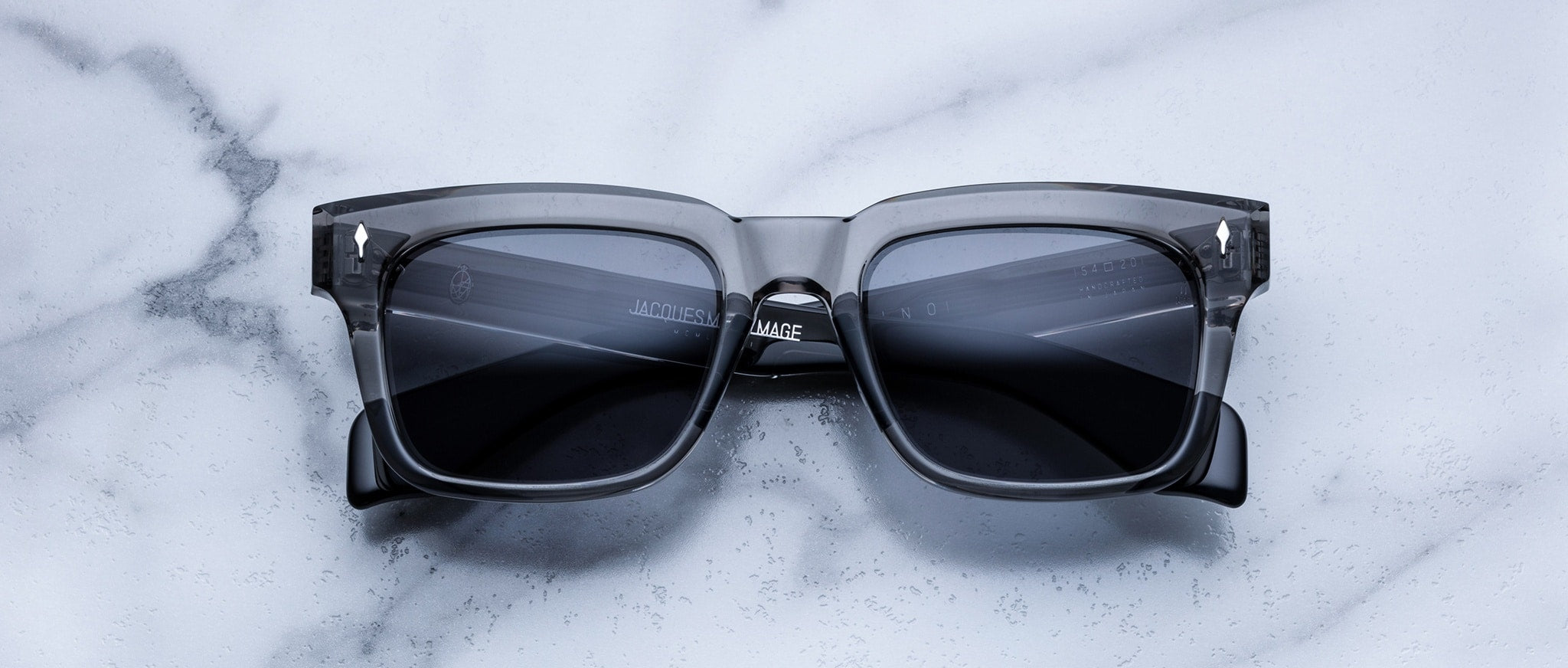 The Jacques Marie Mage Torino features bold rectangular frames in transparent gray handcrafted acetate with dark lenses, displayed on a white marble surface.