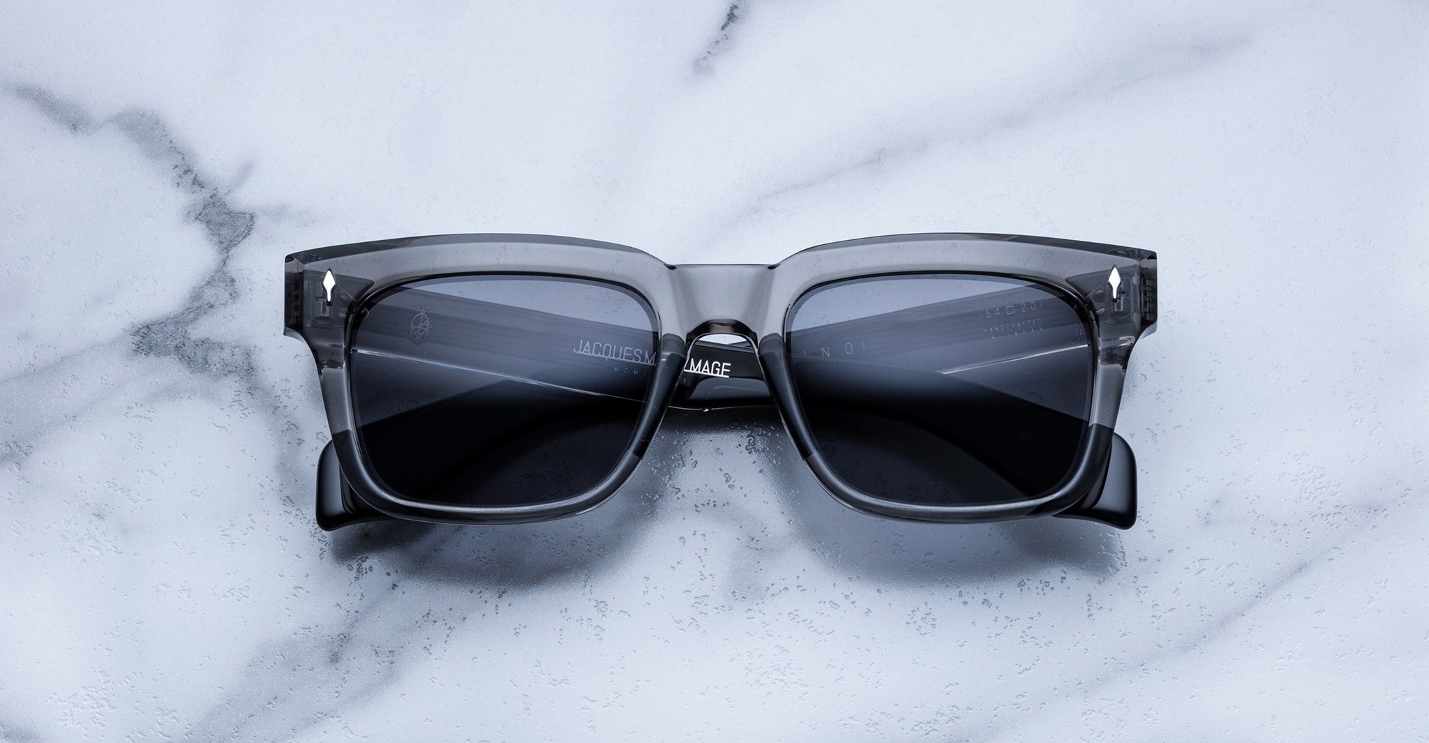 The Jacques Marie Mage Torino features bold rectangular frames in transparent gray handcrafted acetate with dark lenses, displayed on a white marble surface.