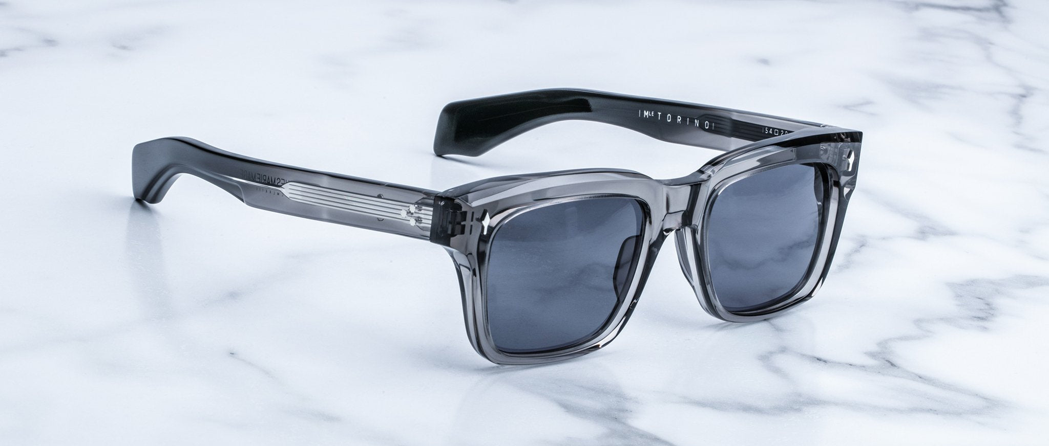 The Jacques Marie Mage Torino sunglasses feature bold rectangular acetate frames and dark square lenses, echoing signature brand style, and are elegantly displayed on a white marble surface.