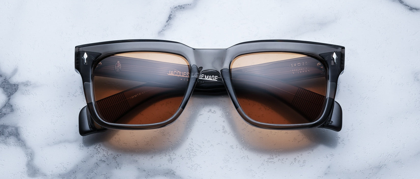 The Jacques Marie Mage Torino eyewear features bold rectangular acetate frames with brown-tinted lenses, showcased on a white marble surface.